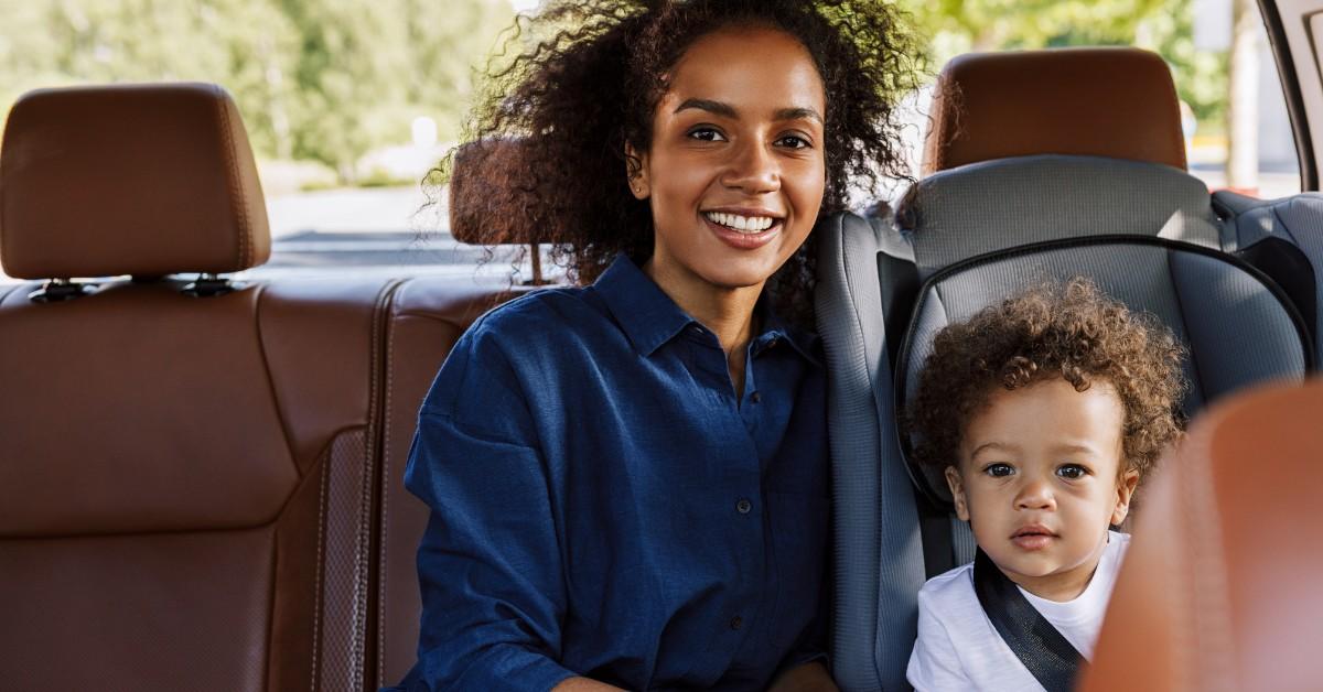 An adult sits next to a child in a car seat in the back of a car